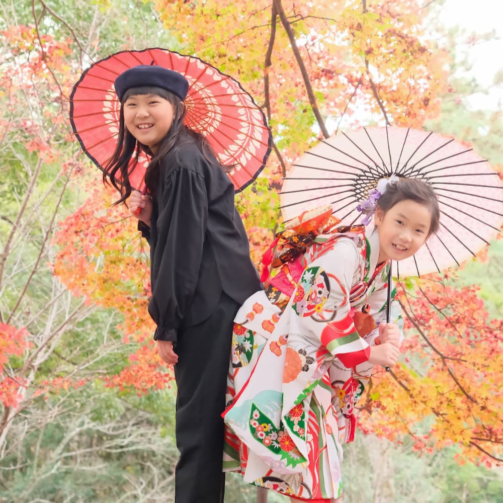 姉と妹で和傘を持って紅葉の下で七五三の記念写真を撮っている
